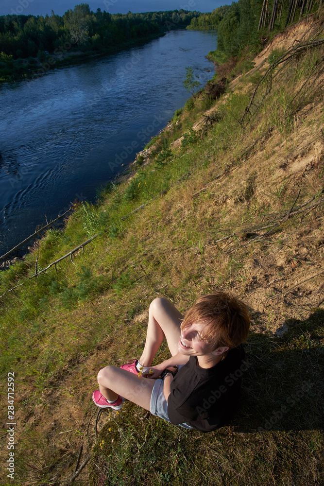 Young caucasian girl sitting on a cliff above forest river and enjoying sunset beams
