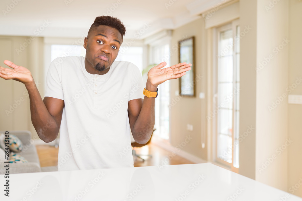Handsome african american man on white table at home clueless and confused expression with arms and hands raised. Doubt concept. Handsome african american man on white table at home clueless and confused expression with arms and hands raised. Doubt concept.