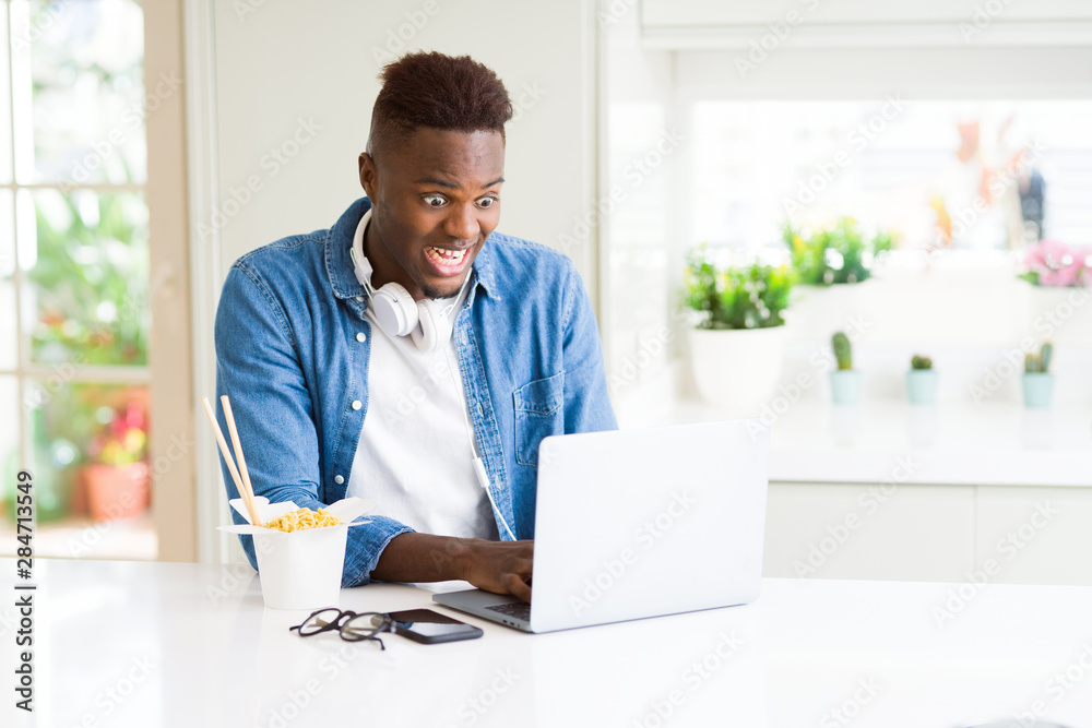 Handsome young african business man eating delivery asian food and working using computer, enjoying noodles smiling Handsome young african business man eating delivery asian food and working using computer, enjoying noodles smiling