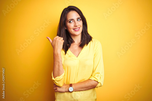 Beautiful elegant woman standing over yellow isolated background smiling with happy face looking and pointing to the side with thumb up.