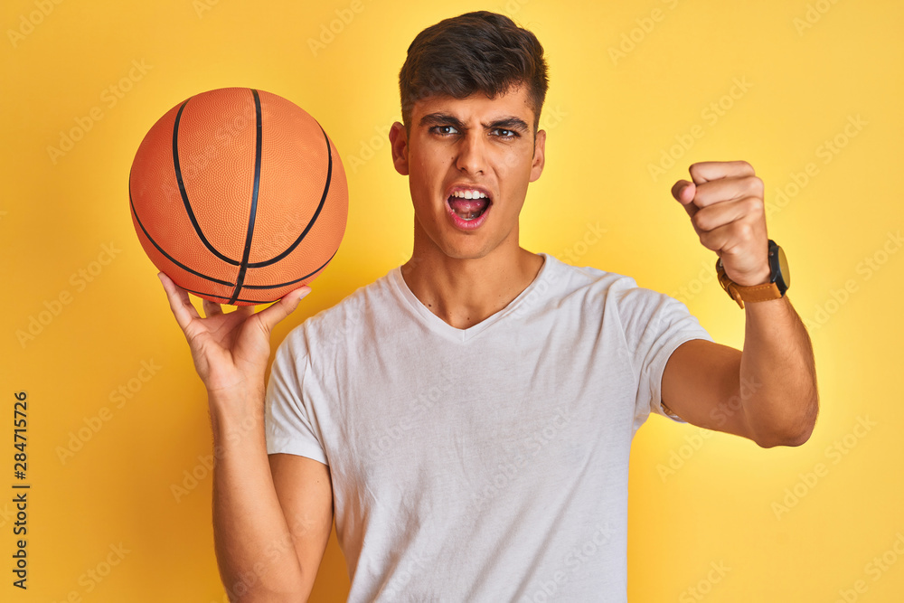 Young indian sportsman holding basketball ball standing over isolated yellow background annoyed and frustrated shouting with anger, crazy and yelling with raised hand, anger concept