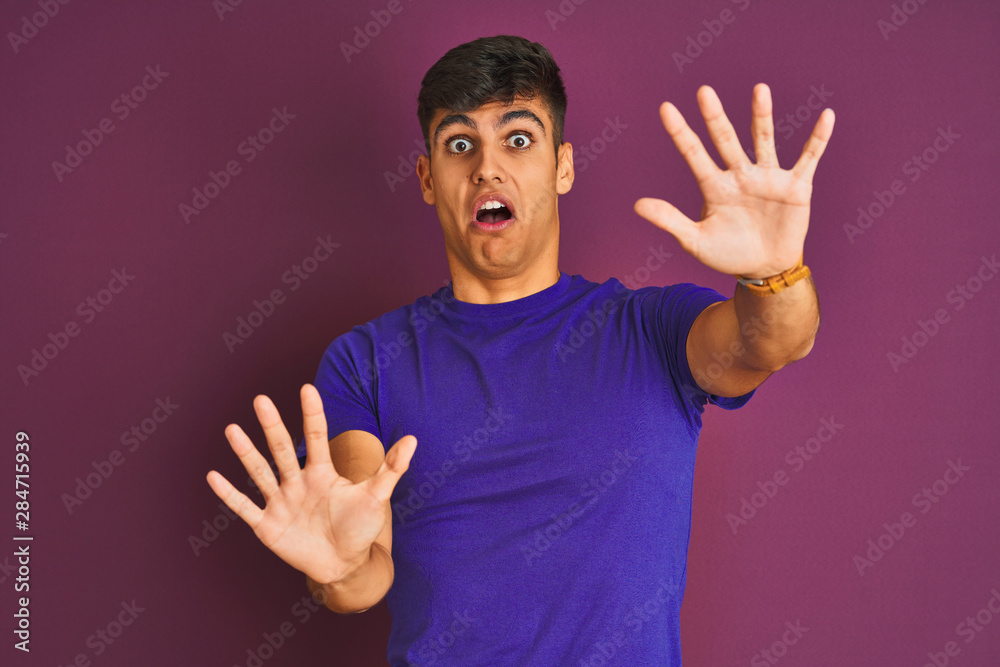 Young indian man wearing t-shirt standing over isolated purple background afraid and terrified with fear expression stop gesture with hands, shouting in shock. Panic concept. Young indian man wearing t-shirt standing over isolated purple background afraid and terrified with fear expression stop gesture with hands, shouting in shock. Panic concept.