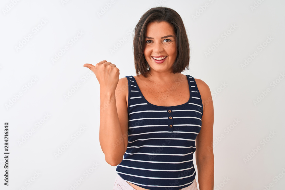 Young beautiful woman wearing blue striped t-shirt standing over isolated white background smiling with happy face looking and pointing to the side with thumb up. Young beautiful woman wearing blue striped t-shirt standing over isolated white background smiling with happy face looking and pointing to the side with thumb up.