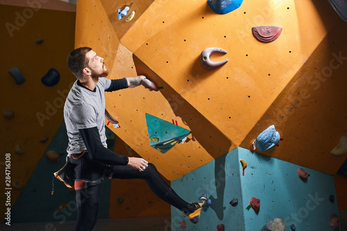 Male physically challenged boulderer standing and staring thoughtfully at difficult climbing wall, planning how to reach the top, dressed in black sportswear, gray t-shirt and special climbing shoes. © alfa27