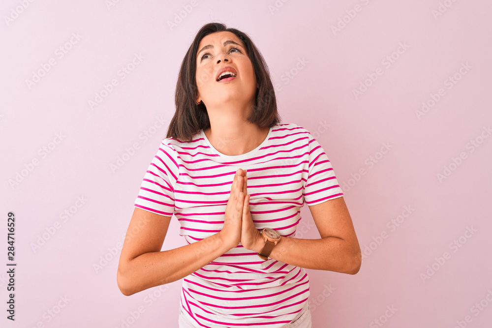Young beautiful woman wearing striped t-shirt standing over isolated pink background begging and praying with hands together with hope expression on face very emotional and worried. Young beautiful woman wearing striped t-shirt standing over isolated pink background begging and praying with hands together with hope expression on face very emotional and worried.