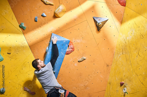 Cropped shot of young strong physically challenged boulderer hanging on large blue artificial rock hold at climbing wall, training indoors. Back view, half-length shot. Climbing sport concept.