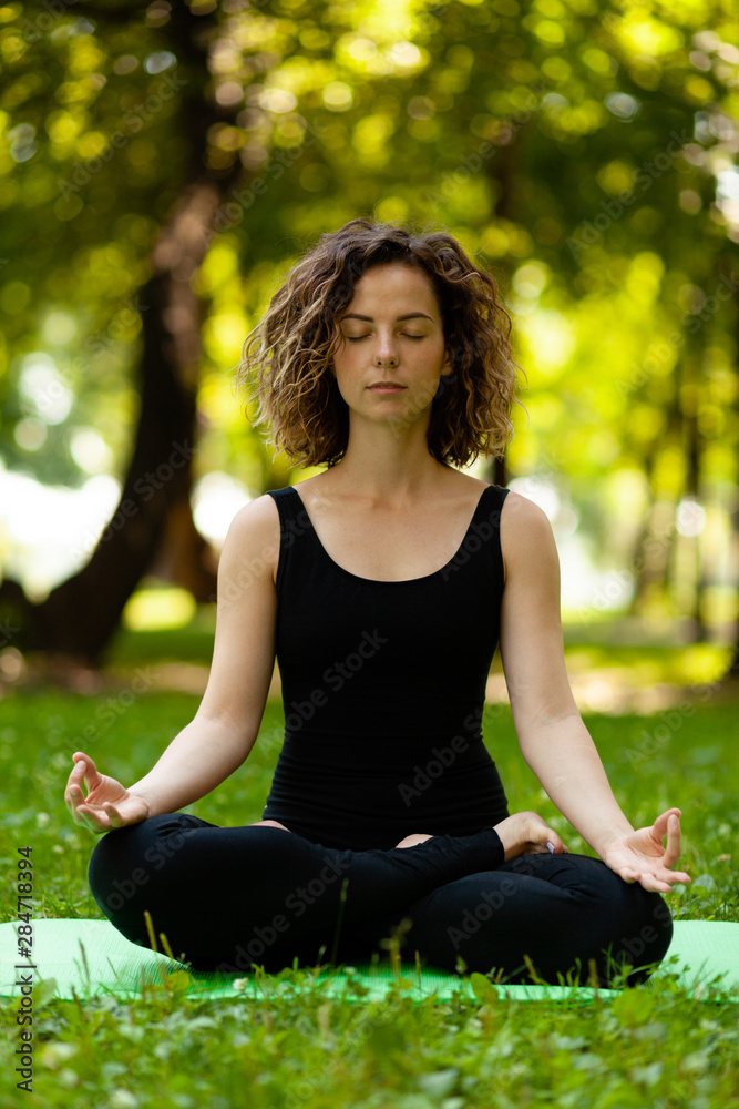 Attractive and tranquil woman sits in lotus position under tree. Yoga and meditation, concentration and relaxation, unity with nature.