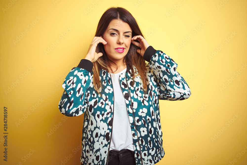 Young beautiful woman wearing casual jacket over yellow isolated background covering ears with fingers with annoyed expression for the noise of loud music. Deaf concept. Young beautiful woman wearing casual jacket over yellow isolated background covering ears with fingers with annoyed expression for the noise of loud music. Deaf concept.