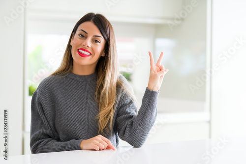Young beautiful woman wearing winter sweater at home smiling with happy face winking at the camera doing victory sign. Number two.