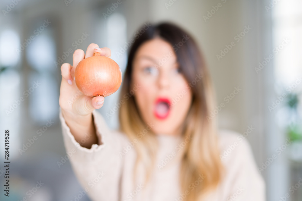Young beautiful woman holding fresh onion at home scared in shock with a surprise face, afraid and excited with fear expression Young beautiful woman holding fresh onion at home scared in shock with a surprise face, afraid and excited with fear expression