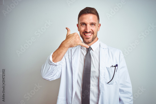 Young handsome doctor man wearing white profressional coat over isolated background smiling doing phone gesture with hand and fingers like talking on the telephone. Communicating concepts. photo