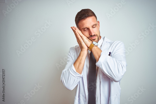 Young handsome doctor man wearing white profressional coat over isolated background sleeping tired dreaming and posing with hands together while smiling with closed eyes. photo