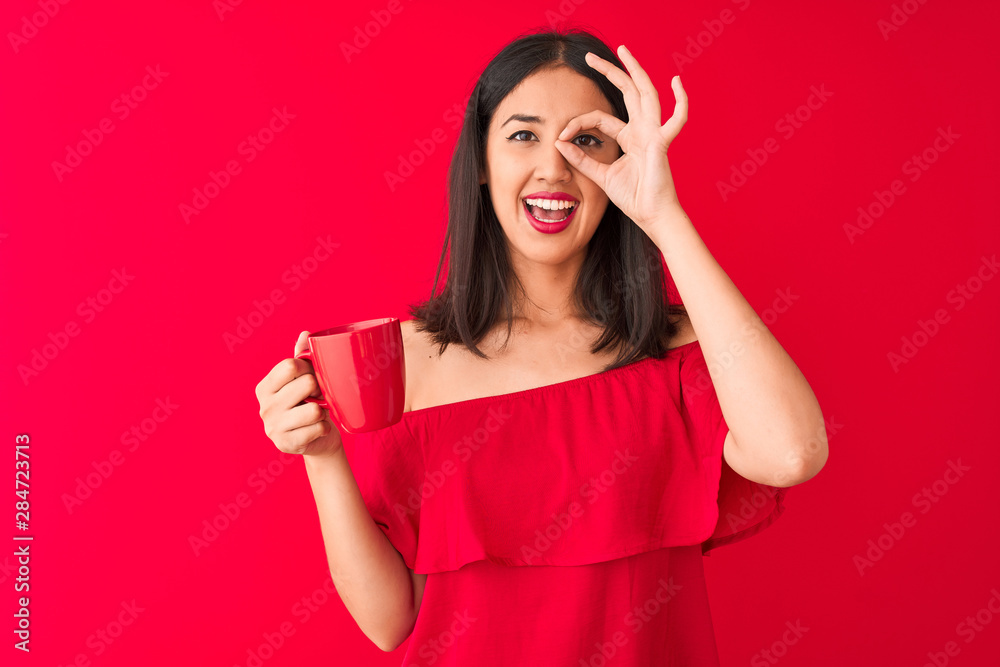 Young beautiful chinese woman drinking cup of coffee standing over isolated red background with happy face smiling doing ok sign with hand on eye looking through fingers Young beautiful chinese woman drinking cup of coffee standing over isolated red background with happy face smiling doing ok sign with hand on eye looking through fingers