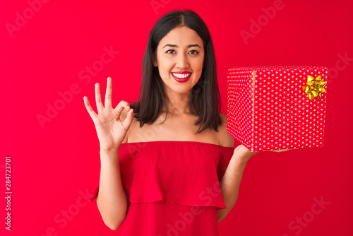 Young beautiful chinese woman holding birthday gift standing over isolated red background doing ok sign with fingers, excellent symbol