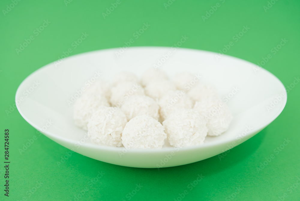 Chocolate covered coconut candies on white plate isolated on green background. Truffle candy set. Selective focus Chocolate covered coconut candies on white plate isolated on green background. Truffle candy set. Selective focus
