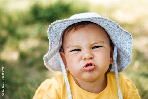 Valokuva Caucasian baby girl is grimacing to the camera in hat and yellow dress