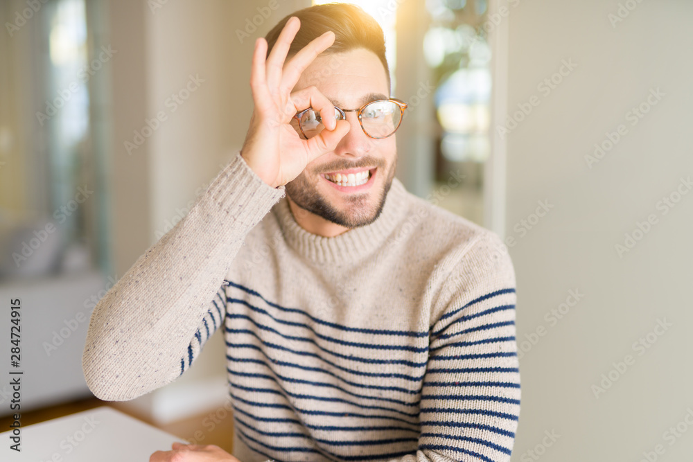 Young handsome man wearing glasses at home doing ok gesture with hand smiling, eye looking through fingers with happy face. Young handsome man wearing glasses at home doing ok gesture with hand smiling, eye looking through fingers with happy face.