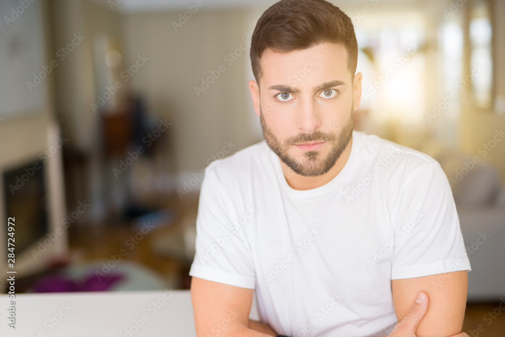 Young handsome man wearing casual white t-shirt at home skeptic and nervous, disapproving expression on face with crossed arms. Negative person. Young handsome man wearing casual white t-shirt at home skeptic and nervous, disapproving expression on face with crossed arms. Negative person.
