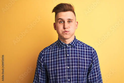Young handsome man wearing casual shirt standing over isolated yellow background puffing cheeks with funny face. Mouth inflated with air, crazy expression.