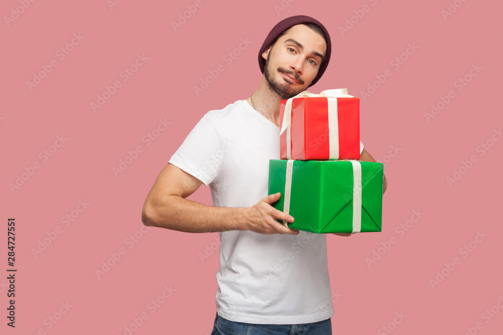 Portrait of handsome bearded young hipster man in white shirt and casual hat standing and holding two present box, looking at camera. Indoor, isolated, studio shot, pink background Portrait of handsome bearded young hipster man in white shirt and casual hat standing and holding two present box, looking at camera. Indoor, isolated, studio shot, pink background