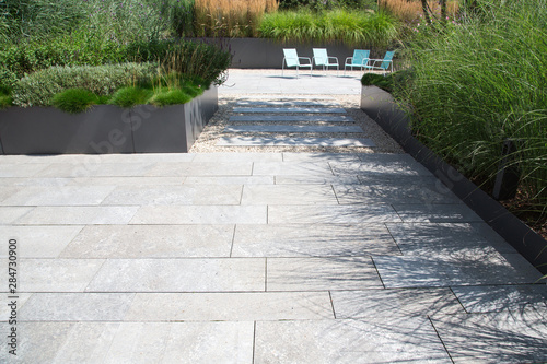 Moderne Garten- und Terrassengestaltung im Materialmix: Terrasse und Gehweg aus Steinplatten umgeben von Schotter und Metall Pflanzgefäßen mit Grünpflanzen