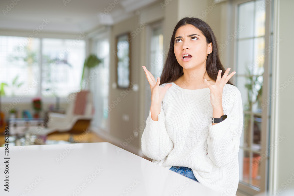 Young beautiful woman at home on white table crazy and mad shouting and yelling with aggressive expression and arms raised. Frustration concept. Young beautiful woman at home on white table crazy and mad shouting and yelling with aggressive expression and arms raised. Frustration concept.