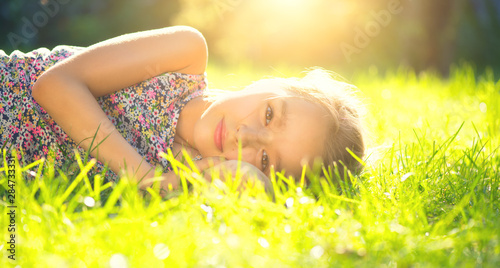 Portrait of a smiling little girl lying on green grass. Cute six years old child enjoying nature in summer park on a lawn