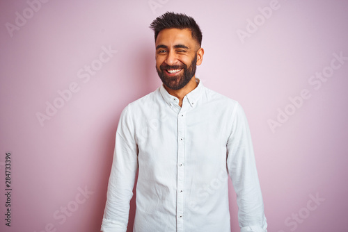Young indian businessman wearing elegant shirt standing over isolated pink background winking looking at the camera with sexy expression, cheerful and happy face.