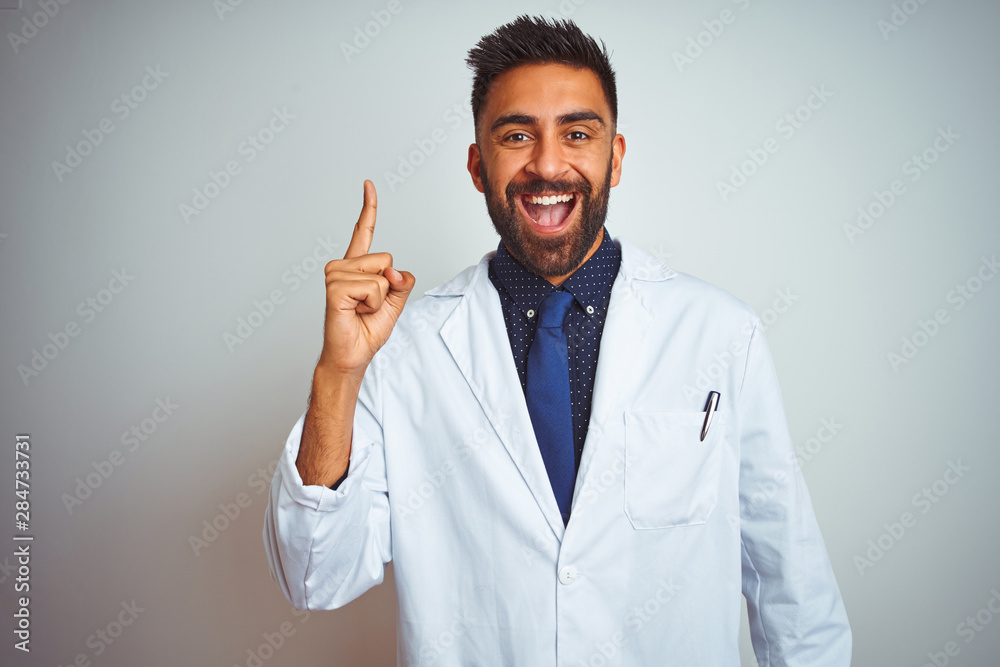 Young indian doctor man standing over isolated white background pointing finger up with successful idea. Exited and happy. Number one. Young indian doctor man standing over isolated white background pointing finger up with successful idea. Exited and happy. Number one.