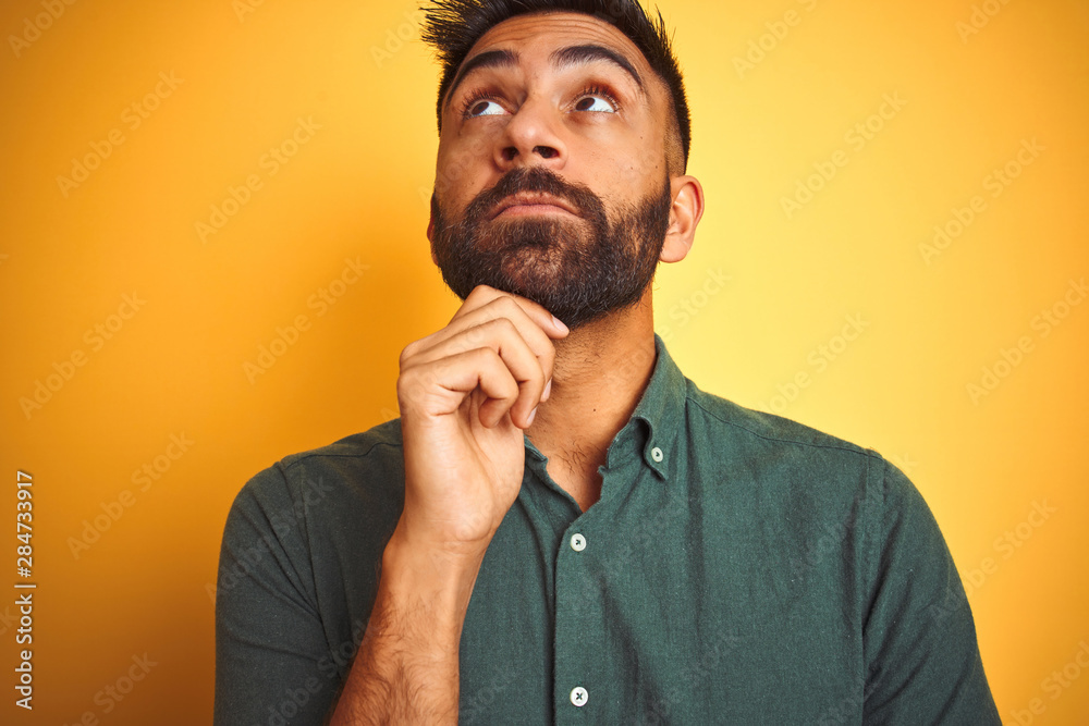 Young indian man wearing green shirt standing over isolated yellow background with hand on chin thinking about question, pensive expression. Smiling with thoughtful face. Doubt concept. Young indian man wearing green shirt standing over isolated yellow background with hand on chin thinking about question, pensive expression. Smiling with thoughtful face. Doubt concept.