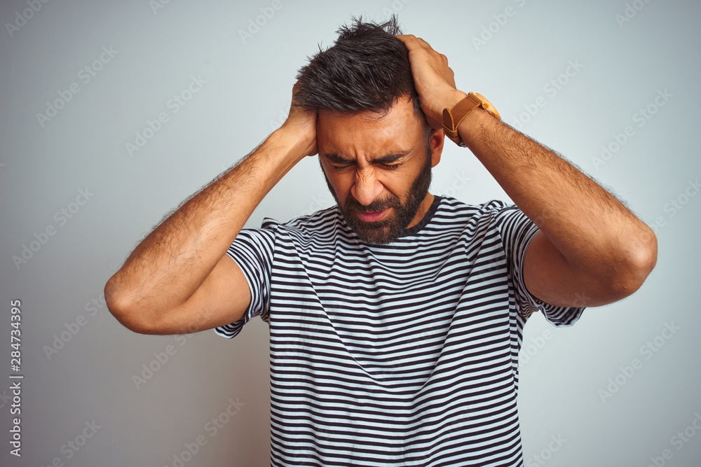 Young indian man wearing black striped t-shirt standing over isolated white background suffering from headache desperate and stressed because pain and migraine. Hands on head. Young indian man wearing black striped t-shirt standing over isolated white background suffering from headache desperate and stressed because pain and migraine. Hands on head.