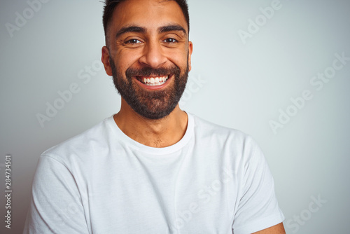 Young indian man wearing t-shirt standing over isolated white background happy face smiling with crossed arms looking at the camera. Positive person.