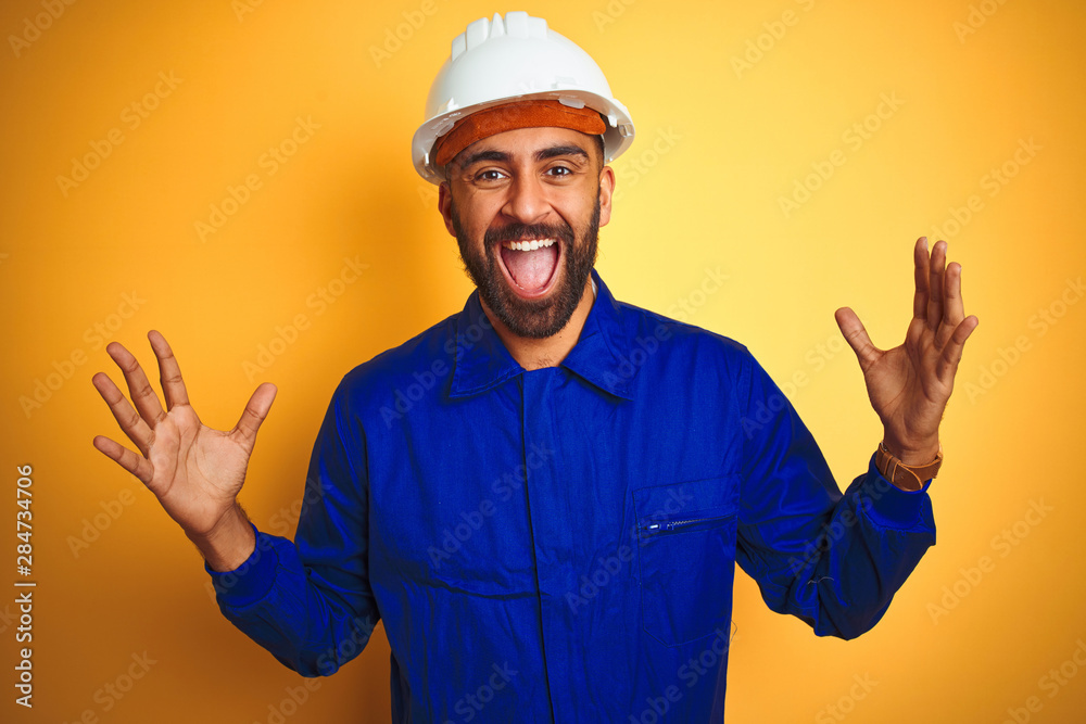 Handsome indian worker man wearing uniform and helmet over isolated yellow background celebrating crazy and amazed for success with arms raised and open eyes screaming excited. Winner concept Handsome indian worker man wearing uniform and helmet over isolated yellow background celebrating crazy and amazed for success with arms raised and open eyes screaming excited. Winner concept