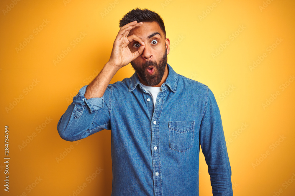 Young indian man wearing denim shirt standing over isolated yellow background doing ok gesture shocked with surprised face, eye looking through fingers. Unbelieving expression.