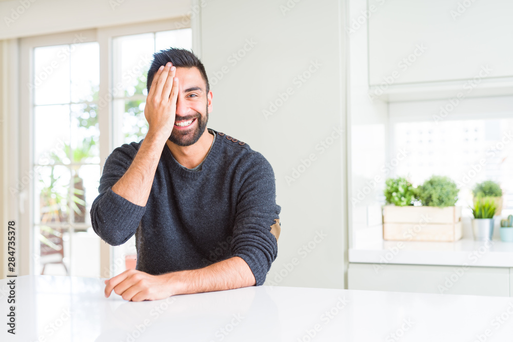 Handsome hispanic man wearing casual sweater at home covering one eye with hand with confident smile on face and surprise emotion. Handsome hispanic man wearing casual sweater at home covering one eye with hand with confident smile on face and surprise emotion.