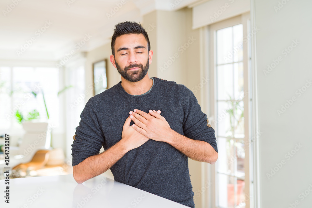 Handsome hispanic man wearing casual sweater at home smiling with hands on chest with closed eyes and grateful gesture on face. Health concept.