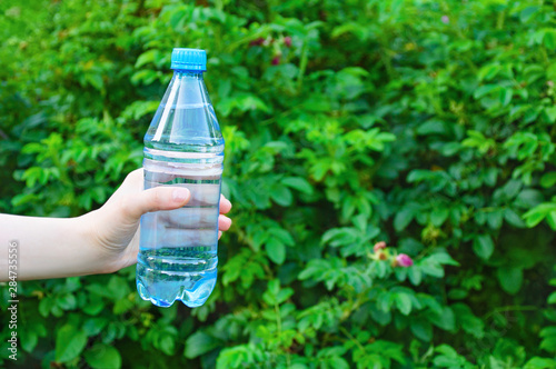 Hand of a caucasian girl holding a water bottle. In the summer on a background of green foliage.