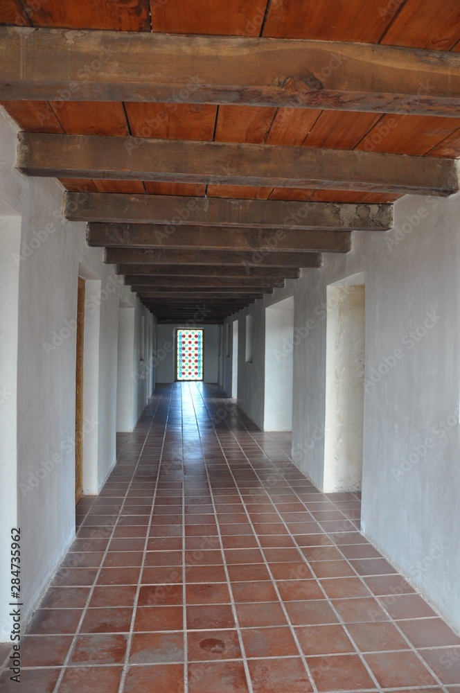 Corridor with white walls, doorways, wooden ceiling and tiled floor, ending with stained glass