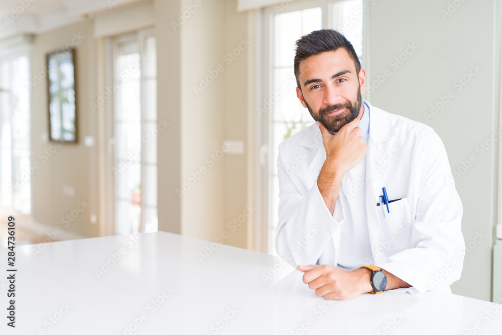 Handsome hispanic doctor or therapist man wearing medical coat at the clinic looking confident at the camera with smile with crossed arms and hand raised on chin. Thinking positive. Handsome hispanic doctor or therapist man wearing medical coat at the clinic looking confident at the camera with smile with crossed arms and hand raised on chin. Thinking positive.