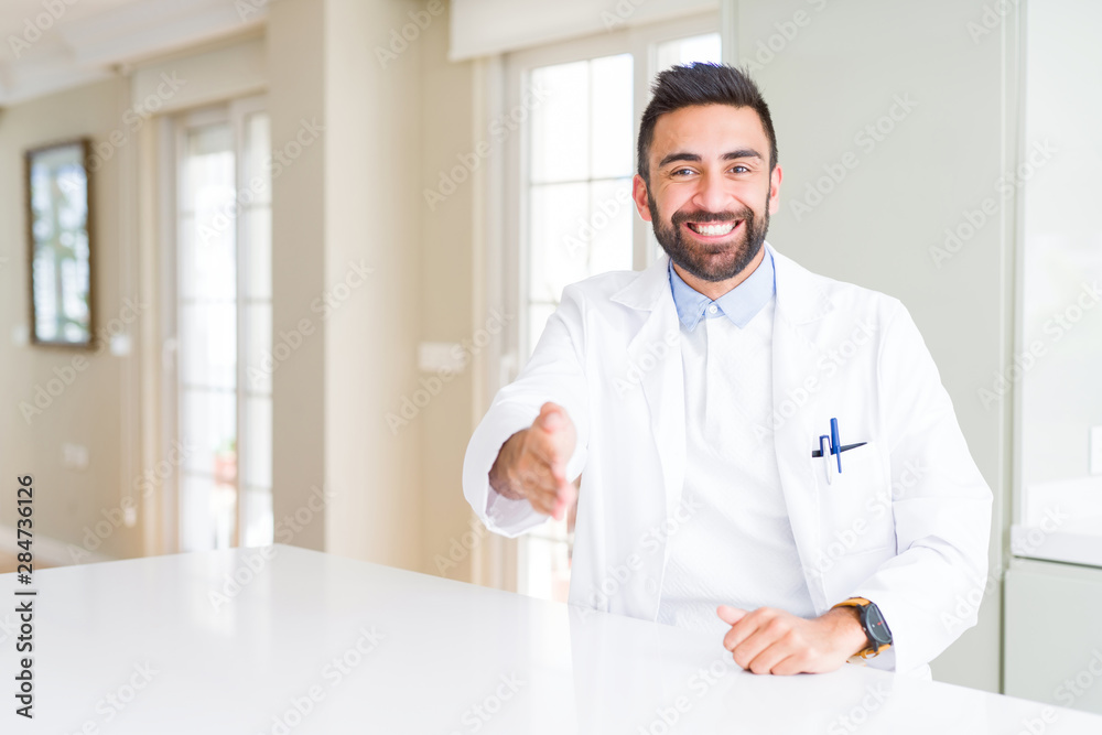 Handsome hispanic doctor or therapist man wearing medical coat at the clinic smiling friendly offering handshake as greeting and welcoming. Successful business.