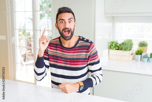 Handsome hispanic man wearing stripes sweater at home pointing finger up with successful idea. Exited and happy. Number one.