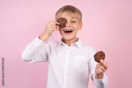 Portrait of confident attractive little boy holds american chocolate chip cookies on the eyse, dressed white stylish shirt clothing isolated pink background photo