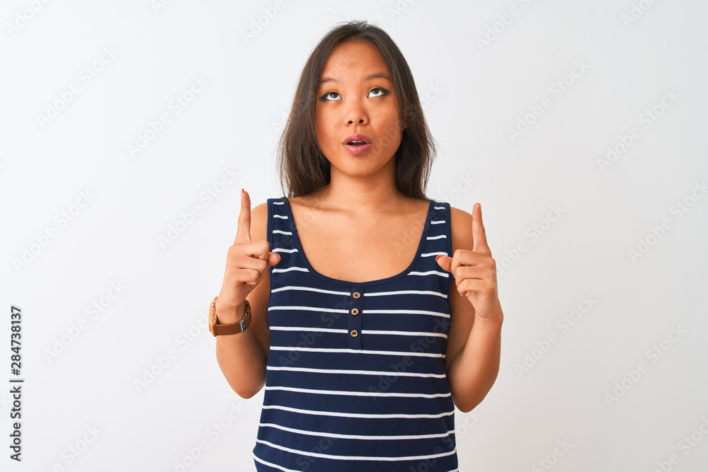 Young chinese woman wearing striped t-shirt standing over isolated white background amazed and surprised looking up and pointing with fingers and raised arms. Young chinese woman wearing striped t-shirt standing over isolated white background amazed and surprised looking up and pointing with fingers and raised arms.