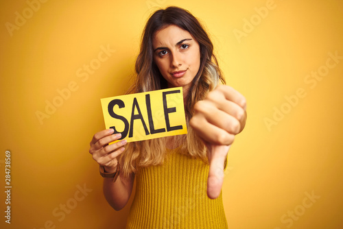 Young beautiful woman holding sale poster over yellow isolated background with angry face, negative sign showing dislike with thumbs down, rejection concept