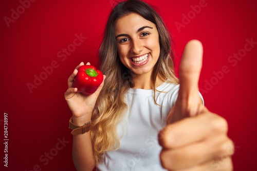 Young beautiful woman holding pepper over red isolated background happy with big smile doing ok sign, thumb up with fingers, excellent sign © Krakenimages.com