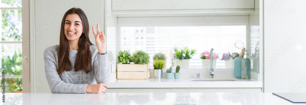 Wide angle picture of beautiful young woman sitting on white table at home smiling with happy face winking at the camera doing victory sign. Number two. Wide angle picture of beautiful young woman sitting on white table at home smiling with happy face winking at the camera doing victory sign. Number two.