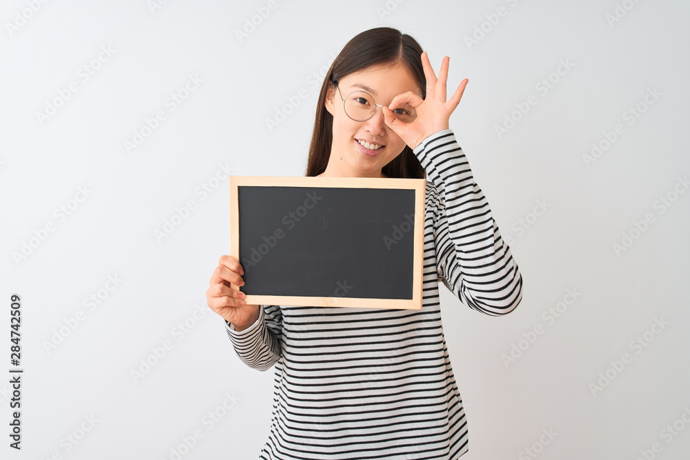 Young chinese woman wearing glasses holding blackboard over isolated white background with happy face smiling doing ok sign with hand on eye looking through fingers Young chinese woman wearing glasses holding blackboard over isolated white background with happy face smiling doing ok sign with hand on eye looking through fingers