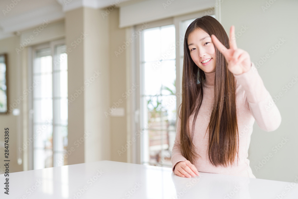 Beautiful Asian woman wearing casual sweater on white table smiling looking to the camera showing fingers doing victory sign. Number two. Beautiful Asian woman wearing casual sweater on white table smiling looking to the camera showing fingers doing victory sign. Number two.