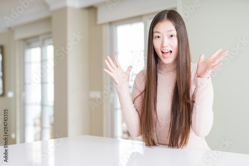 Beautiful Asian woman wearing casual sweater on white table celebrating crazy and amazed for success with arms raised and open eyes screaming excited. Winner concept