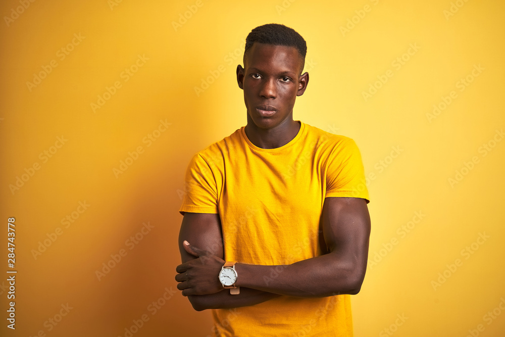 Young african american man wearing casual t-shirt standing over isolated yellow background skeptic and nervous, disapproving expression on face with crossed arms. Negative person. Young african american man wearing casual t-shirt standing over isolated yellow background skeptic and nervous, disapproving expression on face with crossed arms. Negative person.
